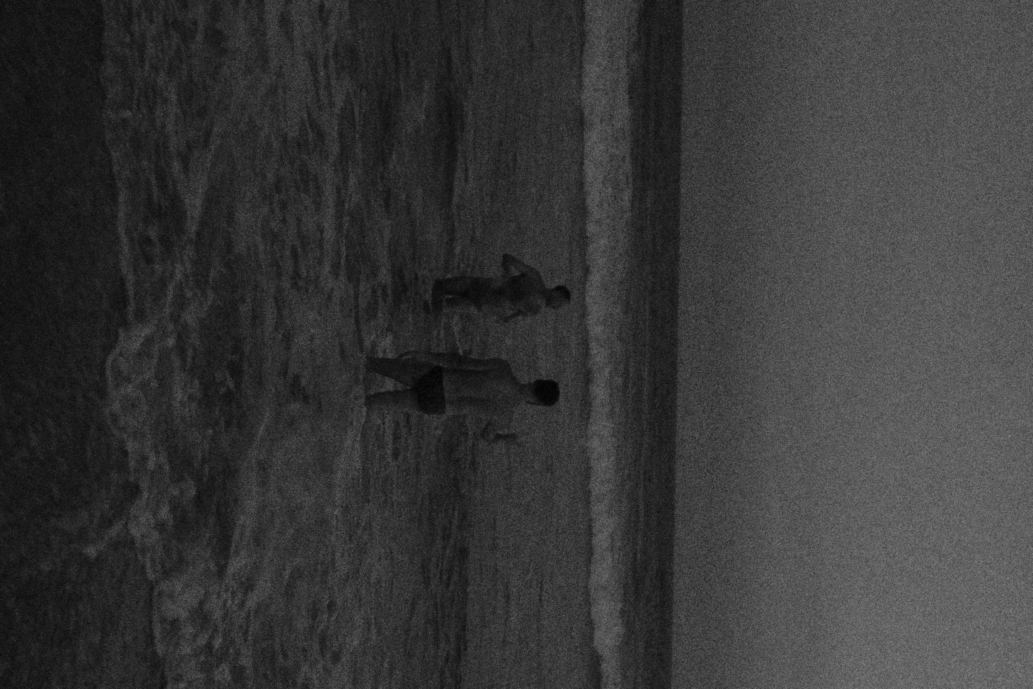 Black and white aerial view of two figures walking along the shoreline at dusk, grainy and atmospheric