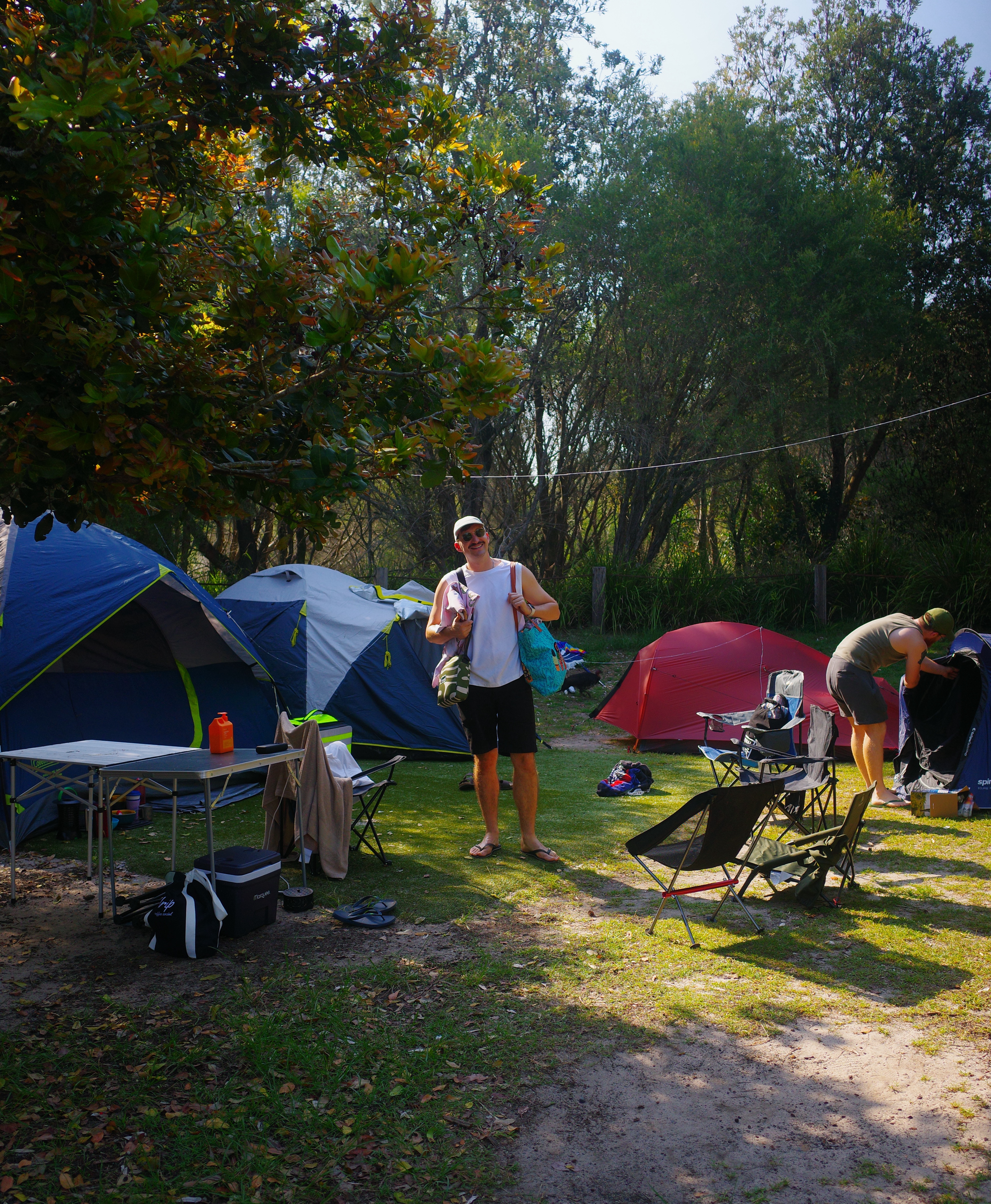 Campsite with blue and red tents among trees, someone standing and smiling with gear in dappled sunlight