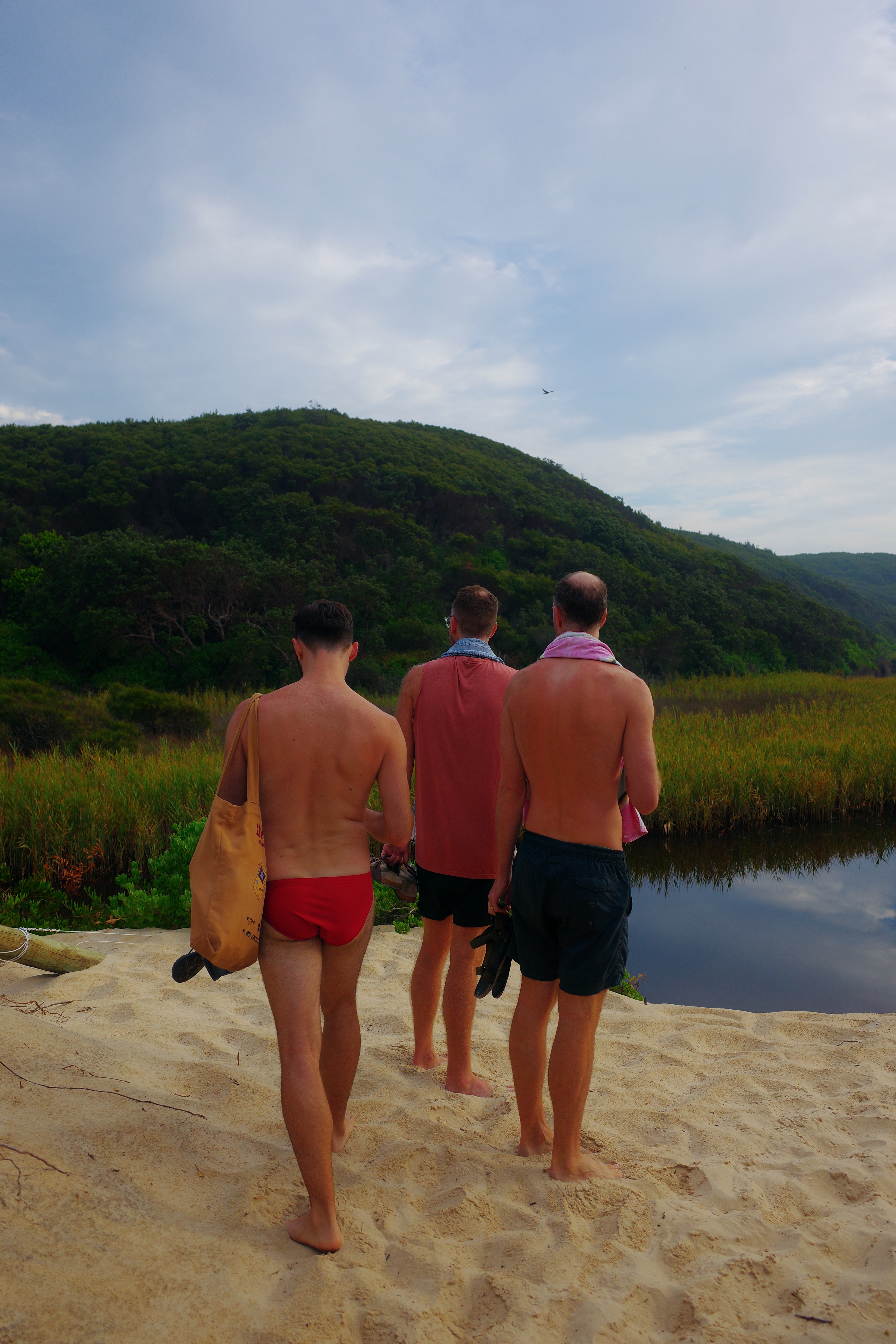 Three friends from behind standing on sand looking out at a still lagoon backed by lush green hills