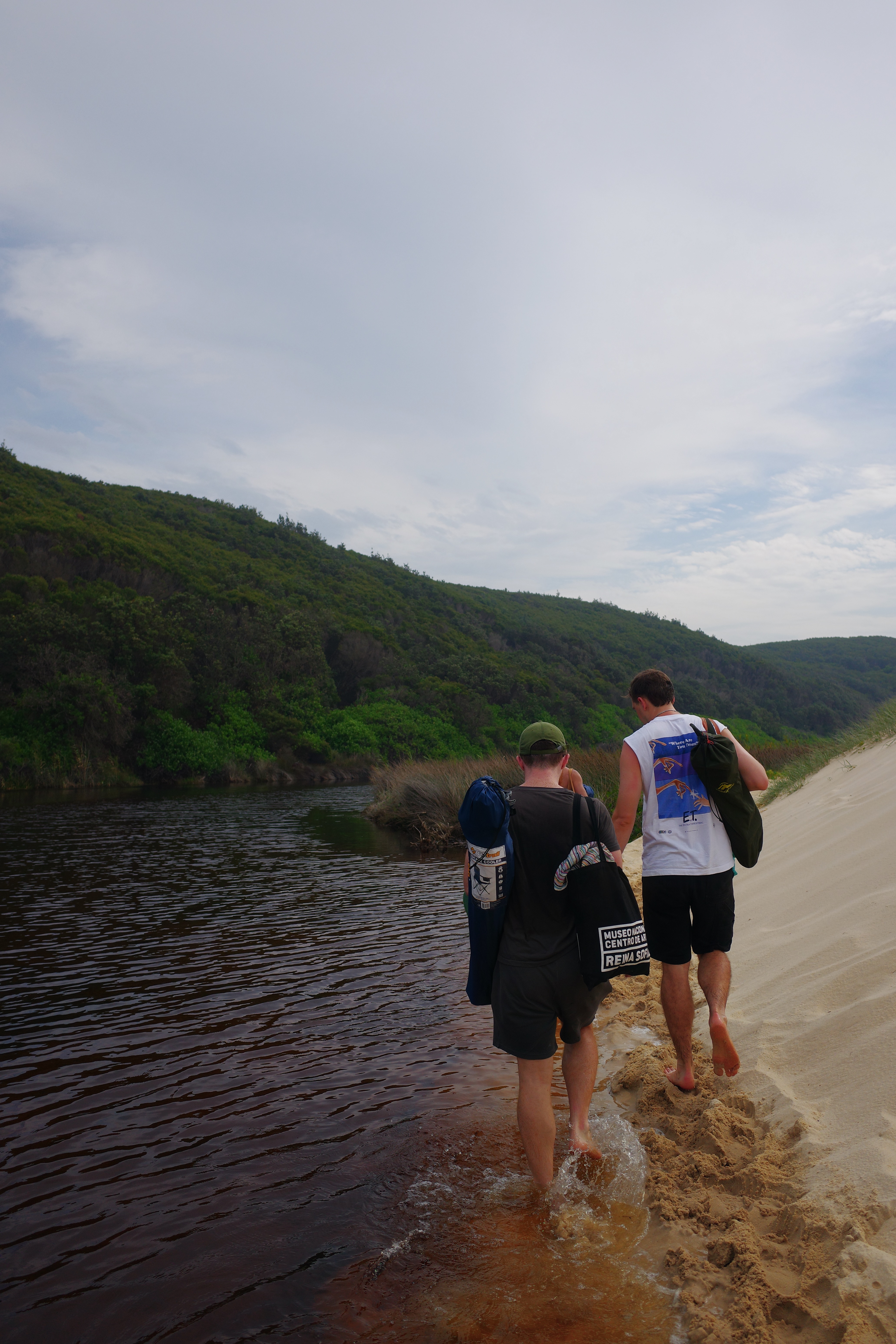 Two people wading through shallow lagoon water carrying bags with green hills rising behind them