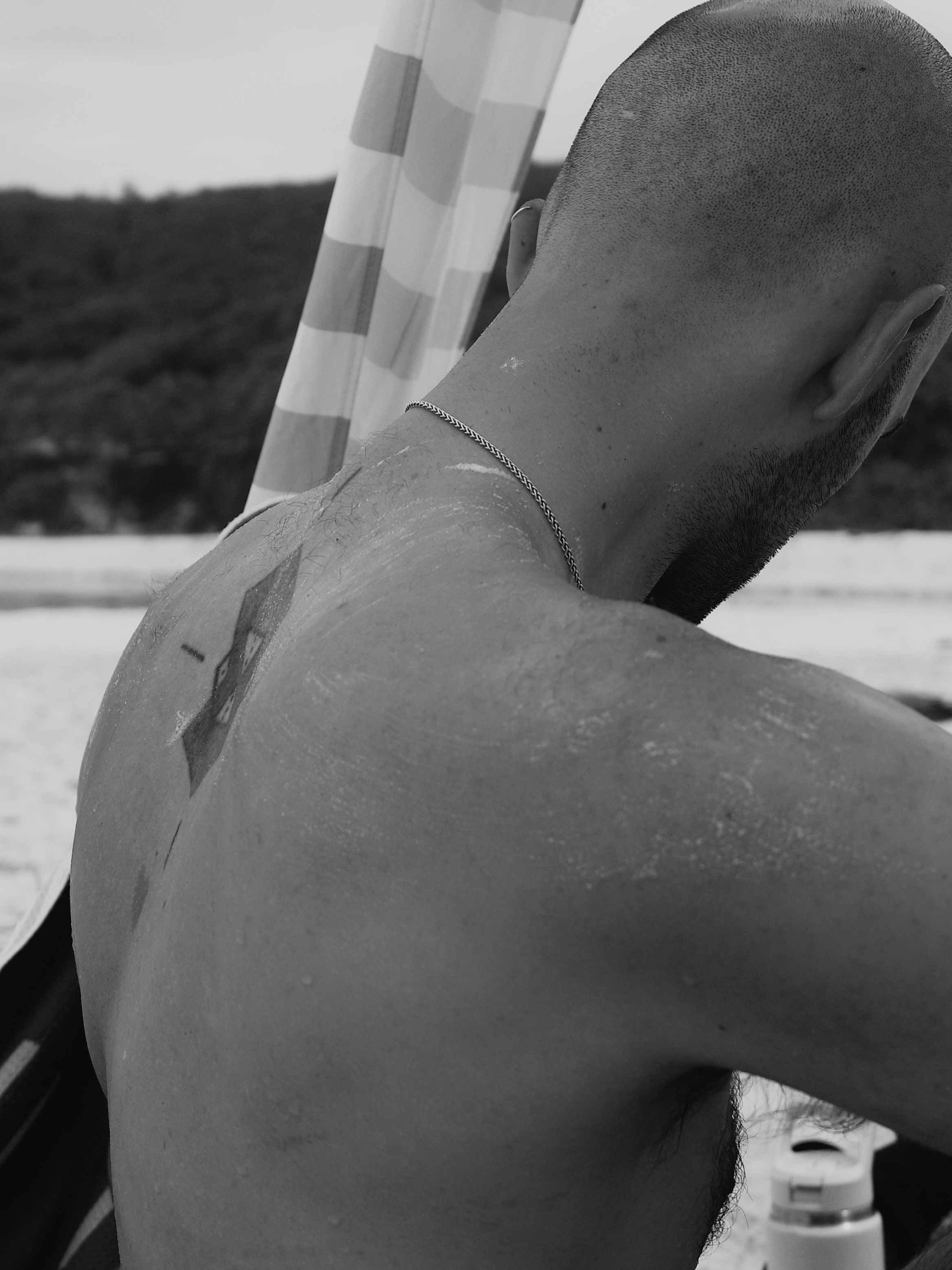 Black and white close-up from behind of someone with sunscreen on their shoulders, chain necklace, and a striped beach umbrella